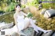 A woman in a white dress sitting on a rock by the water.